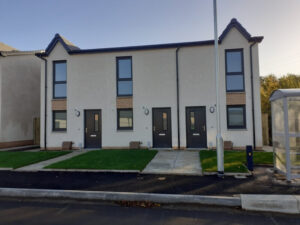 A row of new semi-detached homes with smooth render finish and gray XtremeDoor composite doors, each door accented with contemporary hardware. The houses feature neat, symmetrically placed windows and sandstone accents, set against a backdrop of a freshly landscaped lawn and a clear sky.