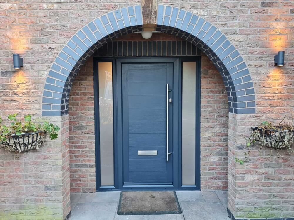 The image showcases a modern grey composite front door set within a red brick arched entryway. Above the door is a semi-circular arch with blue-grey bricks lining its curve. On either side of the doorway are hanging basket planters with greenery, affixed to the brick wall. Two wall-mounted outdoor lanterns cast a warm glow on the bricks. The door features a long vertical handle and a letter slot, suggesting a blend of contemporary design and functionality.
